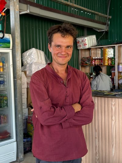The image depicts a man in Phan Son, Vietnam, likely standing inside a small convenience store. He is fair-skinned, with dark hair and eyes, wearing a long-sleeved, maroon shirt and is standing with his arms folded. Behind him, there is a green, corrugated metal wall. In the background, there is another individual with dark hair wearing a striped shirt, partially obscured by shelving filled with various packaged goods. Below the man is a refrigerated display case stocked with food and beverages.