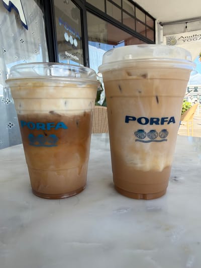 Two iced coffee drinks with cream and ice are on a marble table in front of a building with a large window. The drinks are in clear plastic cups with domed lids. Both cups have the blue logo of "PORFA" with three seashells below it. The building has a black frame around the window and a sign that says "Anutta YOGA". A wicker chair is visible in the background to the right of the drinks, and a yellow chair is also visible further in the background. The scene appears to be outdoors, possibly a cafe or coffee shop. The overall impression is that of a casual, outdoor seating area. The image was taken in Oakland Park, United States of America.