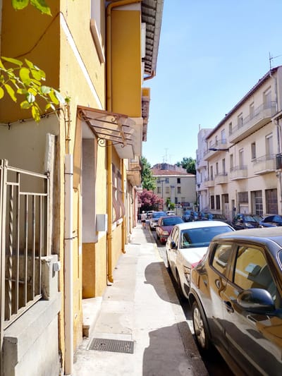 This image depicts a sunlit street scene in Perpignan, France. The perspective is from a narrow sidewalk, looking down the street lined with buildings on both sides.
On the left, a bright yellow building dominates the foreground, with a metal fence and part of a window visible. Green leaves from an overhanging plant cast shadows on the wall. Further down the street, a row of parked cars, mostly sedans and SUVs, are lined up along the curb. The cars are of various colors including black, white, and red.
The buildings lining the street are typical of European architecture, with cream-colored facades, balconies with decorative railings, and shuttered windows. In the background, more buildings are visible, including some with terracotta roofs, and trees with vibrant pink blossoms add a splash of color.
The sky is a clear, bright blue, indicating a sunny day. The shadows cast by the buildings and cars suggest that it is mid-morning or early afternoon. A single person, a woman in dark clothing, is walking away from the camera down the sidewalk on the right side of the street. The overall mood is peaceful and ordinary, capturing a moment of everyday life in a European town. There is no visible text in the image.