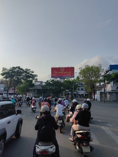This image captures a wide-angle view of a busy urban street scene in Thuận Hóa Ward, Vietnam, likely during late afternoon or early evening due to the soft, warm light and clear sky.
**People:** Approximately 25-30 individuals are visible, primarily operating scooters or motorbikes. Almost all are wearing protective helmets of varying colors, including white, black, beige, and gray. Their attire consists of casual clothing like t-shirts, long-sleeved shirts, and jackets. Most riders are observed individually, while a few appear to have passengers.
**Subjects/Objects:** The foreground and midground are dominated by a dense formation of scooters and motorbikes, positioned in traffic moving away from the viewer. One white SUV, possibly a VinFast VF 8, is partially visible on the far left of the foreground. License plates are discernible on several motorbikes, such as "75-K8 7929" and "75-K1 277.L", with the "75" prefix indicating registration in Thừa Thiên Huế province. In the background, a large red billboard is prominently displayed on a metal frame, featuring the Vietnamese text "Chúc mừng năm mới" (Happy New Year) in gold, accompanied by the national emblem of Vietnam and additional smaller text. Streetlights and power lines are visible.
**Actions/Interactions:** The people are actively engaged in commuting, maneuvering their vehicles in what appears to be a slow-moving or momentarily stopped traffic queue. There is a collective movement in one direction.
**Environment:** The setting is an urban street with an asphalt road surface. Green-leafed trees are scattered along both sides of the road and in the median. Various multi-story buildings line the street; one on the far left has a partially visible sign reading "BẢN HÒA", and another on the far right has a blue facade with white lettering, possibly "AIA". An elevated concrete structure, potentially a bridge or overpass, is visible in the mid-distance. The sky above is clear and light blue with minimal scattered clouds.