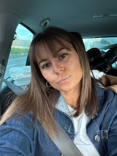 A female individual is depicted in a selfie photograph taken inside a vehicle, likely on a road in or near Bourg-lès-Valence, France. The subject, positioned in the front passenger seat, has medium-brown hair with bangs and shoulder-length straight sections, brown eyes, and a slightly tanned complexion. Her lips are slightly pursed, and she wears a white t-shirt under a blue denim jacket, a gold-colored necklace with a small pendant, and a gold-colored hoop earring in her visible right ear. A gray seatbelt is secured across her chest and lap. Through the left side window of the vehicle, a two-lane road or highway, a light-colored sound barrier or fence, and green foliage are visible under a light blue sky. In the back seat, to the left, a white wrapped item is present, and to the right, dark-colored boots and a section of brown fabric are observed.