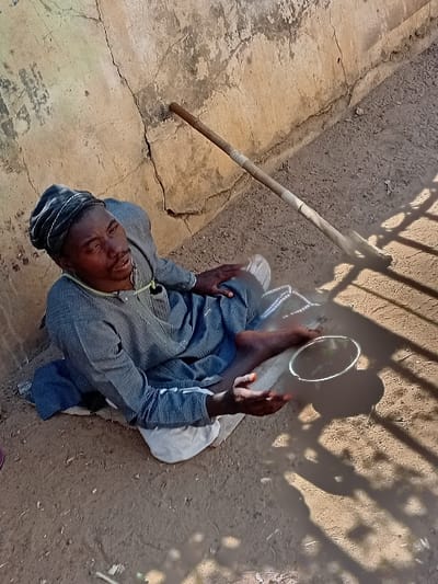 An adult male is seated on dry dirt ground in Kabuga, Nigeria, leaning against a weathered concrete wall. He is wearing a dark skullcap, a long-sleeved dark tunic, and a lighter fabric beneath him. A light-colored cord hangs around his neck. His legs are bent, and his feet are bare. He holds a small, dark metallic bowl in his right hand, extended forward. A light brown wooden stick, wrapped with lighter material, rests on the ground beside him, leaning against the wall. The scene is illuminated by strong sunlight, casting distinct grid-like shadows on the ground and the bowl. The wall shows cracks and discoloration.