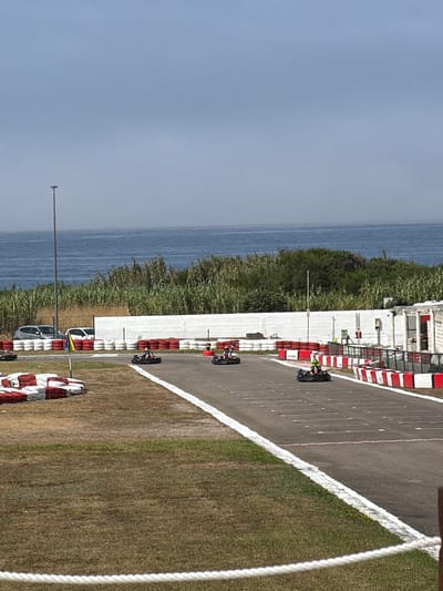The image depicts a go-kart track in Matosinhos, Portugal, situated near the Atlantic Ocean. The track is asphalt, bordered by white and red tires and safety barriers. Several go-karts are visible on the track, with drivers wearing helmets and racing gear. In the background, lush green vegetation lines the coast, with the ocean stretching to the horizon under a hazy sky. A street lamp is visible on the left side of the frame, and some cars are parked in the distance behind the tire barriers. The scene appears to be a sunny day, possibly in the late morning or early afternoon.