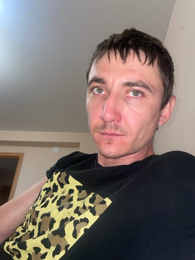 This is a medium close-up, head-and-shoulders selfie taken by a man indoors. The man has light skin, short dark brown hair, and green eyes. He is wearing a black t-shirt with a gold and black leopard print design. He has a light mustache and stubble. The background is out of focus and appears to be a room with a light-colored wall and a ceiling. The lighting is soft and appears to be coming from above and to the left. There is a doorway visible in the background on the left. The image was taken in Laishevo, Russia.