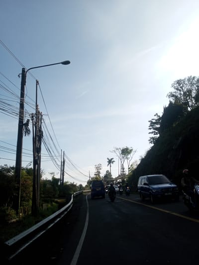 The image depicts a road scene in Phou Khoun, Laos. The road is asphalt with a white dividing line, and a guardrail is present on the left side of the frame. A blue Isuzu Panther car and several motorcycles are visible, indicating vehicular traffic. The surrounding environment features utility poles laden with wires, trees, and a hillside with dense vegetation to the right. The sky is light blue, and the overall lighting suggests daytime conditions.