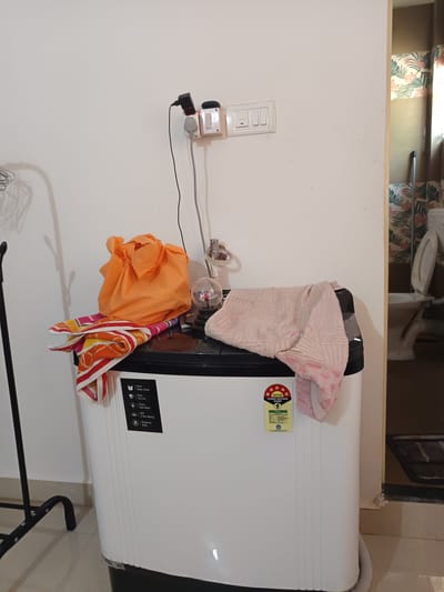 The image shows a top-load washing machine in a bathroom. On top of the washing machine are an orange cloth bag and two towels, one folded and the other draped. To the left of the washing machine is a black metal clothing rack. On the wall above the washing machine is a light switch with two outlets and a power adapter plugged into one. To the right of the washing machine, a toilet is visible in the background, with its lid up. The washing machine has a BEE 5-star rating sticker on its front. The scene appears to be indoors, in a residential setting. There is no specific indication of the location being in Oxel, India.