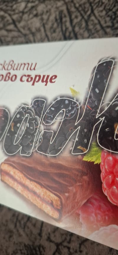 The image shows a close-up of a chocolate-covered confectionery product, possibly a wafer or cake, with a layer of filling. The product is decorated with images of blackberries and a raspberry. The text "ckBumu" and "ovo сърце" is visible in a reddish-brown font on a white background, suggesting it is a brand name or product description in Bulgarian. The product is placed on a textured, brown surface that resembles fabric or a tablecloth. The background is out of focus, showing a patterned surface. The scene is an indoor still life, with no people or specific environmental context related to Kaolinovo, Bulgaria.