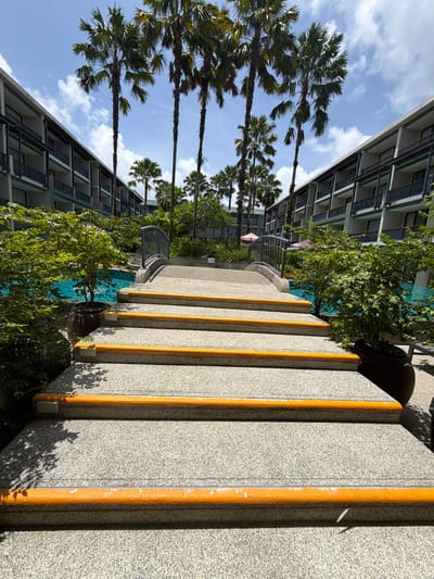 In Bang Muang, Thailand, a sunlit resort scene unfolds with a wide-angle perspective. The foreground features a set of prominent concrete stairs with bright orange non-slip treads, leading upwards. Flanking the stairs are lush green potted plants and shadowy foliage. Beyond the stairs, a tranquil blue swimming pool is visible, crossed by a decorative pedestrian bridge with a dark gray railing. Tall palm trees tower over the scene, with their fronds silhouetted against a bright blue sky dotted with white clouds. On either side of the pool and bridge, multi-story hotel buildings with balconies and dark railings rise up, creating a symmetrical architectural frame for the tropical landscape. The overall impression is one of a well-maintained resort environment on a clear, sunny day.