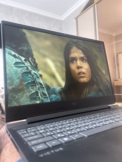 A close-up, angled shot shows a black laptop with the screen displaying a still image of a woman with dirty skin, wide eyes, and slightly open mouth. The image suggests she is in a state of distress or shock. The background of the screen image is blurry and appears to be a forest or natural setting. The laptop's keyboard is visible in the foreground. The laptop appears to be an HP Victus laptop. The top left corner of the screen also displays performance metrics like FPS, Frame Time, GPU Util, GPU Power, GPU Temp (42°C), and CPU Util. The location Topolovgrad, Bulgaria is not relevant as it cannot be determined from the image alone.