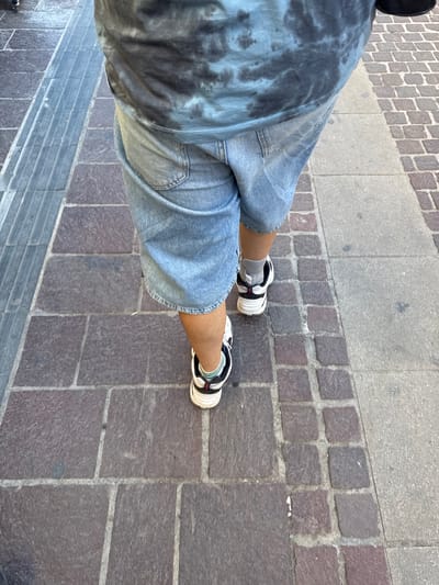 A person walks on a sidewalk in Chania, Greece. The sidewalk is paved with rectangular, reddish-brown stones arranged in a pattern. To the left of the paved section is a narrower strip of gray concrete with raised, ridged lines, likely a tactile paving system for the visually impaired. The person is wearing a tie-dye t-shirt with blue and black patterns, light blue denim shorts, gray socks, and black and white athletic shoes with a red accent stripe. The image is taken from a low angle, looking down at the person's legs and feet as they walk away from the camera. The lighting suggests it is daytime, with shadows visible on the ground.