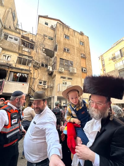 An outdoor scene in Bnei Brak, Israel, depicts a group of individuals in the foreground against the backdrop of a severely damaged multi-story residential building.
The central background features a dilapidated, light-brown concrete apartment building showing significant structural compromise. A large section of its facade on the upper left appears to have collapsed or been blown out, exposing interior elements, debris, and tangled wires. Numerous windows are shattered or missing, with some frames bent. Air conditioning units are visibly mounted on the exterior walls, some askew. To the right, another building, lighter in color, is partially visible. The sky overhead is clear and light blue.
In the mid-ground and foreground, a crowd of people, predominantly men, are gathered. From left to right:
1. A man in a dark kippah and an orange reflective vest, bearing Hebrew text "בני ברק" (Bnei Brak) and an emblem, stands talking to another man. He has a dark beard.
2. A man wearing a dark olive-green fedora-style hat, a white long-sleeved shirt, and eyeglasses smiles while looking towards the left, extending his left arm slightly. He has a dark beard.
3. On the right, a man with a large, black fur shtreimel (hat), a dark coat, and a long grey beard carries a small child on his left hip. He wears eyeglasses and looks downwards.
4. The child, a young boy with reddish-blonde hair and a straw hat, wears a yellow plaid shirt and blue overalls. He holds a red plush toy, possibly a chicken, with colorful accents. The child looks off to the left with a neutral expression.
Other individuals are visible in the background, including one person wearing an emblem resembling a red cross. The overall impression is one of observation in the aftermath of an event, given the extensive damage to the building.