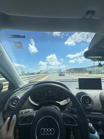 The image shows the interior of a car, looking out through the windshield at a highway on a sunny day with a blue sky and white clouds. The dashboard is visible, with a steering wheel, speedometer showing 100 km/h, and a central display screen. The driver's left hand, with light-colored nail polish, is on the steering wheel. Outside the car, other vehicles are driving on the highway, and there are overhead structures and buildings in the distance. The scene appears to be on a highway, possibly in or near a city, with a modern infrastructure visible. Without further information, it's not possible to pinpoint the exact location in German, Bulgaria.