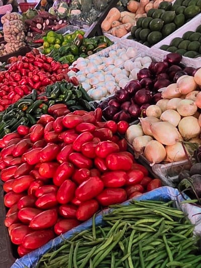 The image displays a vibrant market stall in Voluntari, Romania, laden with a diverse assortment of fresh produce. In the foreground, a large pile of bright red, Roma-type tomatoes dominates the view, some appearing bruised or slightly imperfect. Adjacent to the tomatoes, a generous bin of long, slender green beans fills the bottom right quadrant of the image. Behind the tomatoes, a collection of what appear to be red chilies or small red bell peppers is visible. Further back, a variety of other vegetables are displayed, including white onions, red onions, green peppers, and what look like avocados. The stall is organized with produce arranged in bins or piles, suggesting a typical open-air market setting. The lighting suggests it is daytime, likely outdoors. There are no people visible in the frame.