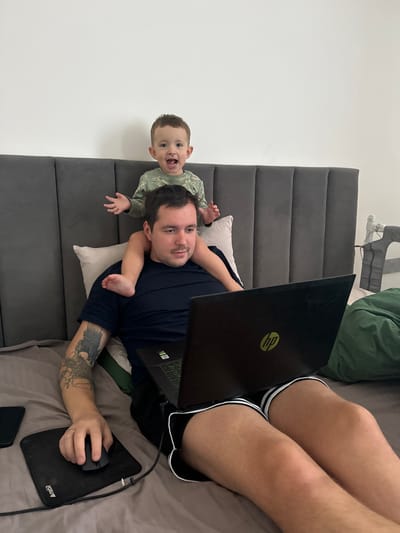 An adult male and a young child are depicted indoors in what appears to be a bedroom in Селекционер-2 Снт, Russia. The adult male, with short dark hair and a tattoo on his left forearm, wears a dark blue t-shirt and black shorts with white trim. He is reclining on a bed with grey bedding, operating a black HP laptop positioned on his lap, and a black wired mouse on a black mousepad to his left. A dark smartphone rests next to the mousepad. A light-colored pillow supports his head against a dark grey upholstered headboard with vertical panels. A male toddler, with light brown hair and wearing a green long-sleeved shirt with a pattern, is seated on the adult's shoulders, with his legs resting on the adult's upper back. The child has his arms raised and spread slightly, and his mouth is open in an expression of excitement. The background consists of a plain white wall. To the right, a dark green fabric item and a white electronic device are partially visible.
