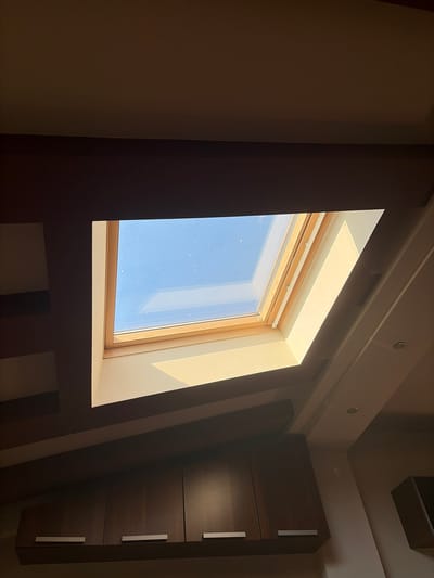 The image shows an interior view looking upwards at a skylight on a ceiling. The skylight is rectangular and framed with light-colored wood. Through the skylight, a bright blue sky with some small white specks, possibly dust or faint clouds, is visible. Light streams through the skylight, illuminating a portion of the ceiling and casting shadows. Below the skylight, on the ceiling, there are recessed panels and what appear to be light fixtures. Further down, a row of dark brown cabinets with silver handles is mounted on the wall. The overall impression is a modern interior with natural light flooding in from above. The location is identified as Vladaya, Bulgaria.