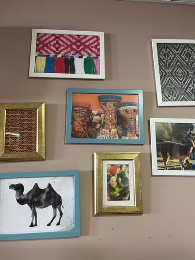 The image shows a wall with six framed pictures. The wall is painted a light brown color. The top left picture is a white frame with a picture of colorful woven fabric. The bottom left picture is a blue frame with a black and white silhouette of a camel. The top right picture is a white frame with a black and white patterned image. The picture directly below this frame is a white frame containing a picture of a llama. The picture below the top left picture is a light blue frame with an orange backdrop and three doll-like figures with colorful clothing. The last picture on the bottom right is a gold frame with a picture of a food dish. This appears to be a wall in a home or business located in Las Palmas de Gran Canaria, Spain at coordinates: latitude: 28.146, longitude: -15.43. The pictures are hung with visible white nail holes, though the placement is otherwise orderly and aesthetically pleasing. The pictures appear to be of diverse origin and theme, including traditional clothing, animals, food, and folk art.