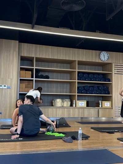 The image depicts an indoor yoga studio with wooden paneling and shelves. In the foreground, a man in a black t-shirt and shorts is seated on a yoga mat, holding a small black ball. A woman in a maroon top is to his left, also on a mat, smiling. Behind them, a woman in a white t-shirt is reaching for a dark ball on a shelf. The shelves contain yoga blocks, rolled-up yoga mats, and storage bins labeled "CLEAN" and "USED". A clock is visible on the wall above the shelves. The floor is wooden, and several yoga mats are laid out. The overall environment suggests a class or practice session.