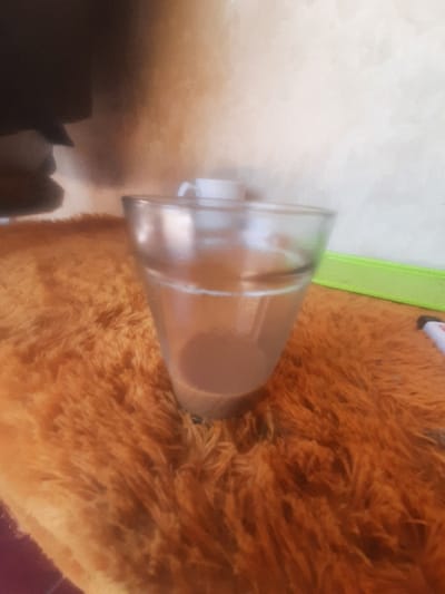 The scene depicts an indoor environment in Bandar Lampung, Indonesia. A transparent glass, approximately two-thirds full of a light brown liquid, likely coffee or tea, is centrally positioned on a textured, shaggy orange-brown surface, resembling a rug or blanket. In the background, an off-white and light brown wall is visible. To the upper right of the glass, a white ceramic cup is partially observed. Further to the right, a bright green horizontal strip or object is present, and a small white object with a red tip, possibly a marker or lighter, is partially visible at the far right. No individuals are present in the frame, and no specific actions or interactions are depicted.