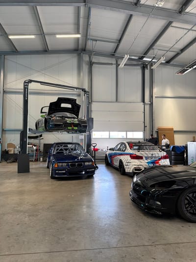 The image displays an interior view of a commercial automotive workshop located in Meuspath, Germany. The facility features an industrial design with high ceilings, exposed metal structural elements, and a light-colored corrugated metal roof. Overhead, multiple linear fluorescent light fixtures provide illumination. The floor is composed of grey concrete.
In the left-center, a black "Twinbusch.de" branded two-post car lift is elevated, holding a dark green and black race car, resembling a BMW M3 E46 GTR, with its hood open and the engine bay exposed. Directly beneath the elevated vehicle, a dark blue BMW E36-series sedan, extensively modified for racing with a visible internal roll cage, is parked on the floor.
To the right, a white BMW M3 (F80-series) race car with prominent blue, red, and black livery, including "TUNING" and "PAUL CARE" branding, is parked facing away from the viewer. Its rear displays a large aerodynamic wing and diffuser, and a German license plate "BM:WP 9991" is affixed. Two adult males are visible near this vehicle; one in a dark shirt, the other in a white long-sleeved shirt, appearing to be observing or interacting with the car or its immediate surroundings. Behind them, a stack of black tires and other workshop equipment are visible.
In the lower right foreground, the front portion of a black sports car, consistent with a Chevrolet Corvette (C5 or C6 generation) due to its distinct headlight design and low-slung fascia, is partially visible. The background wall features horizontal windows and sections of large overhead garage doors, suggesting multiple entry points. The scene depicts a functional environment for high-performance automotive maintenance and preparation.