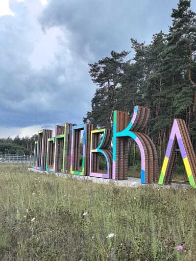 In the city of Razumnoye, Russia, large, colorful, three-dimensional letters stand in a field of tall, dry grass. The letters spell out "РАЗУМНОЕ", which translates to "Razumnoye". Each letter is constructed from a metallic framework with a weathered, brown finish, and features colorful geometric patterns in shades of purple, pink, blue, green, yellow, and orange on the inside. Behind the letters, a dense forest of pine trees stretches across the horizon. To the left, a highway is visible with a silver car driving on it and a sound barrier along the edge. The sky is overcast with dramatic, dark gray clouds. The overall scene depicts a vibrant art installation set against a natural, somewhat muted background.
