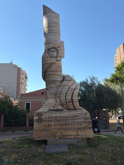 The image depicts a monument featuring a carved face on a tall, textured stone structure. The monument is located in L'Hospitalet de Llobregat, Spain. Inscriptions are visible on the base of the monument, including what appears to be a coat of arms and text that reads "A RAFAEL CASANOVA / L'ESPERIT DE / L'11 DE SETEMBRE DE 1714". The surrounding environment includes a clear blue sky, buildings in the background, trees, and a paved area with a rainbow-colored stripe. A person is walking in the background on the right side of the image, and a parked scooter is visible behind the monument. The ground in front of the monument is covered with grass and scattered pieces of colorful confetti.