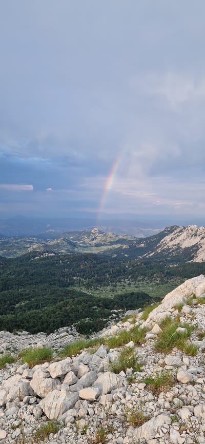An outdoor, high-angle, vertical, full shot captures a scenic mountainous landscape under a cloudy sky, with a faint rainbow visible. The foreground is dominated by a rocky, uneven terrain covered in scattered grey rocks and patches of green grass. Beyond the immediate foreground, the terrain slopes down into a valley filled with dense, dark green forest. Further in the distance, rolling hills and mountains are visible, characterized by lighter rocky outcrops and vegetation. The sky is a pale blue, with scattered grey clouds, some of which appear heavy and dark, suggesting recent or impending rain. The rainbow, a subtle arc of colors, appears in the middle distance, emerging from behind a dark cloud and arching across the sky. The overall scene depicts a natural, rugged environment, possibly in or near Ubla, Bosnia and Herzegovina, at a time when the weather is transitional. There are no people or discernible structures in the image.