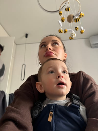 This image captures an indoor scene in Gribovo, Russia, featuring two primary subjects and a partial third, observed from a low angle looking upwards. A female adult occupies the mid-ground, her head tilted back, eyes focused upwards and slightly right, with a subtly pursed mouth and dark hair, wearing a brown long-sleeved garment and small hoop earrings. She is holding a young child, approximately a toddler, who is positioned in the foreground, facing the viewer but also looking directly upwards with an open mouth, revealing light-colored eyes and prominent drool on the lower lip and chin. The child is dressed in a grey knitted sweater with a patterned upper chest area, covered by dark blue overalls with black buckle straps and a central zipper featuring a tan, leather-like pull tab.

Directly overhead, suspended from the white ceiling, is a decorative mobile consisting of a golden hoop from which approximately ten to twelve yellow and black striped crocheted bees with white wings hang on white strings, encircling a central, light-brown spherical object. To the left of the adult's head, a black pendant light fixture descends from the ceiling. On the upper right, a white wall-mounted air conditioning unit is visible against the plain white wall. A blurred figure, possibly male, wearing a light-colored garment with a dark design, is partially visible on the far left edge of the frame, behind the adult. The overall environment is brightly lit, consistent with a domestic or childcare setting.