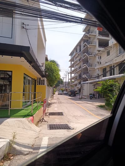 The image, taken from inside a vehicle, shows a street in Hua Hin City Municipality, Thailand. On the left is a yellow and white building with a black awning and artificial grass. A red safety barrier is placed along the sidewalk. The street has a yellow dividing line and several drainage grates. On the right is a tall, white building with exterior staircases. Several vehicles, including a black pickup truck and a scooter, are visible further down the street. Overhead, there are numerous tangled electrical wires. The sky is light blue.