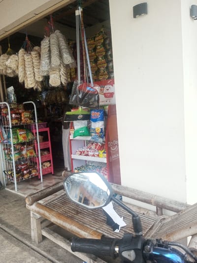 A small shop sells a variety of snacks and goods. Hanging from the storefront are strings of packaged crackers. Inside, shelves display chips, cookies, and other packaged items. Brooms lean against the wall. A wooden bench sits in front of the shop, with the handlebars and mirror of a blue motorcycle visible. Two light fixtures are mounted on the wall to the right of the shop entrance.