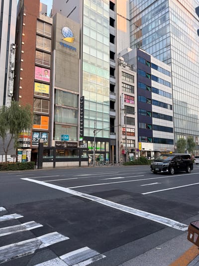 The image captures a street scene in Chuo, Japan, likely during the daytime, given the illumination. In the foreground, a pedestrian crossing with white stripes is visible on the asphalt road. A black minivan is driving in the right lane of the road, with its license plate showing "46-53". The background is dominated by a row of multi-story buildings with a mix of modern and traditional architectural styles. Some buildings feature large glass facades, reflecting the surrounding environment. Signs and advertisements are visible on the buildings, indicating various businesses such as a "BOOK CAFE," a "Total Body Care" center, and other commercial establishments with Japanese text. A green tree with sparse foliage stands to the left of the buildings, and a lamppost is also present. The sky is overcast, suggesting diffused lighting conditions. The scene depicts a typical urban environment with traffic, buildings, and commercial activity.