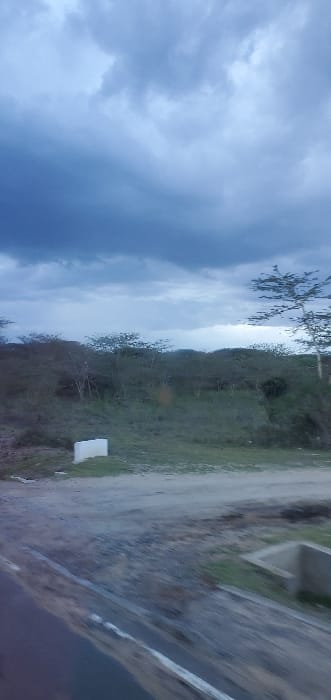 The scene, set in Mua ward, Kenya, depicts an outdoor environment under a heavily overcast sky with dense, dark grey clouds. In the foreground, a paved road section, exhibiting motion blur, occupies the lower-left. Adjacent to it, a lighter-colored dirt road or path runs horizontally. A rectangular, white concrete marker is situated at the edge of the dirt path, slightly left of center. To the far right, a concrete drainage culvert or structure is partially visible. The midground and background feature undulating terrain densely covered with trees and shrubs, many appearing leafless or with sparse foliage. No human individuals, animals, or active subjects are discernible within the frame.