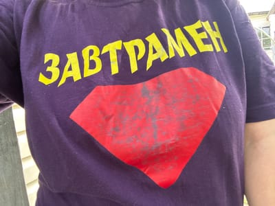 A person wearing a purple t-shirt with a yellow Cyrillic text and a red diamond-shaped emblem is pictured outdoors. The text on the t-shirt appears to read "ЗАВТРАМЕН" (Zavtramen). The red emblem has a distressed or faded appearance with greyish markings. The background shows siding of a building, possibly a house, and a white railing or fence. The lighting suggests it is daytime. The scene is not specifically located in the city of Ленинградская, Russia.