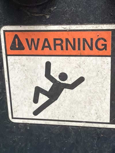 A warning sign is affixed to a dark, textured surface. The sign has an orange header with a black exclamation point inside a triangle and the word "WARNING" in black block letters. Below the header, a black pictogram depicts a person falling. The pictogram shows a figure with arms and legs outstretched as if in mid-fall. The background of the pictogram area is a mottled white. The sign appears to be made of metal or a durable plastic and shows signs of wear, including some scuffs and dirt. The surrounding dark surface appears to be part of a piece of machinery or equipment. This image was taken in Murfreesboro, United States of America.