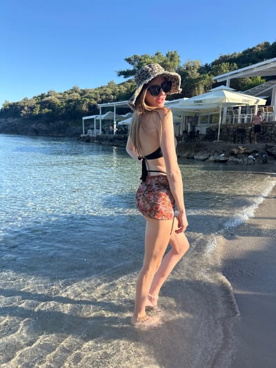 A young woman stands in the shallow, clear water at the edge of a sandy beach in Kubasi, Montenegro, turning her head back over her right shoulder to face the camera. She wears a leopard-print bucket hat, dark sunglasses, a black bikini top, and a fitted, patterned mini-skirt featuring floral and leopard motifs. A white beaded anklet is visible on her left ankle, submerged in the water. Her long, blonde hair flows down her back. The woman's lips are slightly parted in a faint smile. Behind her, calm, clear water stretches out, reflecting sunlight. The background features a steep, tree-covered hillside adorned with white, open-air structures, including sun umbrellas branded with "TUBORG," indicative of a beachside bar or restaurant. Tables and chairs are visible under these structures. A bare-chested man in dark swim trunks stands near the structures on the right. The sky above is clear blue.