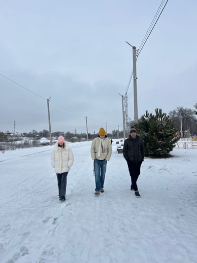 Three individuals walk on a snow-covered path in Synelnykove, Ukraine, on an overcast winter day. The ground is uniformly covered in packed snow with visible footprints. From left to right: a person in a pink beanie, white puffer jacket, and dark pants; a person in a yellow beanie, light-colored hooded jacket with a "DIESEL" logo, a white scarf, and blue jeans; and a person in a dark beanie, dark olive winter jacket, and dark pants. All three are walking towards the camera. To the left, a line of concrete utility poles with overhead wires recedes into the distance, with sparse residential buildings and bare trees beyond. To the right, another line of utility poles is visible, followed by a dense evergreen shrub in the mid-ground. Further right and in the background, elements of a children's playground with yellow metal structures are discernible, with one small figure present near the playground.