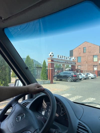 The image shows a view from inside a car, looking through the windshield. The driver's hand is on the steering wheel, which has a Mercedes-Benz logo. The dashboard instruments are visible, indicating the car is likely in motion. Outside, a brick building with the text "КАРПАТИ" and "СПОРТКОМПЛЕКС им. Лена банка" is visible. Two soccer balls are placed on poles in front of the building, one larger and one smaller. There are parked cars and some greenery. The scene appears to be during the daytime with clear skies. The text on the building suggests a sports complex named "Carpathians" named after Lena Bank. The image has a slight tilt.
