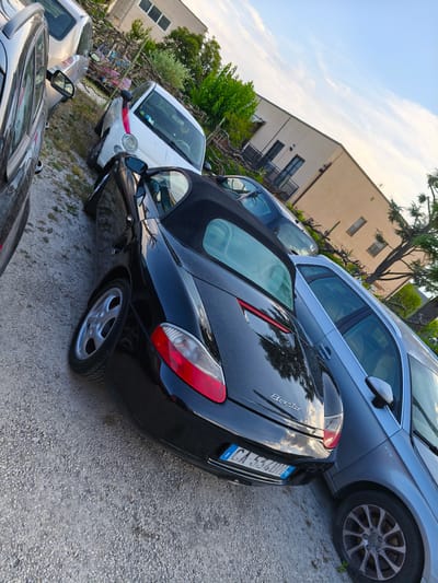 In Chiugiana, Italy, a black convertible sports car, identified as a Porsche Boxster by its badging, is parked on a gravel surface. The car is angled, with its rear facing the viewer and its front partially obscured by another vehicle. To the left of the Boxster, a portion of a dark-colored car is visible. To the right and behind the Boxster, a silver sedan is parked. In the background, a light-colored residential building with balconies and windows is situated. Lush green trees and foliage surround the area, and a bright, partly cloudy sky is overhead. The scene appears to be a parking area, likely in a residential or semi-rural setting.