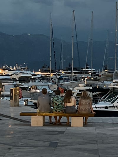 In Kumbor, Montenegro, at dusk, a group of people are seated on a bench facing a marina filled with sailboats. The sky is a deep blue, indicating the transition from day to night. The boats, with their tall masts, are moored together, creating a dense, organized pattern. Some lights are visible on the boats, suggesting that the marina is active even as darkness falls.
The people are casually dressed and appear relaxed, enjoying the evening atmosphere by the water. One person is wearing a patterned dress, another a light-colored top, and two others are wearing casual shirts. They are sitting close together, perhaps a family or group of friends. The overall mood seems peaceful and contemplative, as they observe the marina. The paved walkway and the textured wall behind them suggest an urban or developed waterfront area. The image captures a serene moment in a popular tourist destination, highlighting the beauty of the marina at twilight.