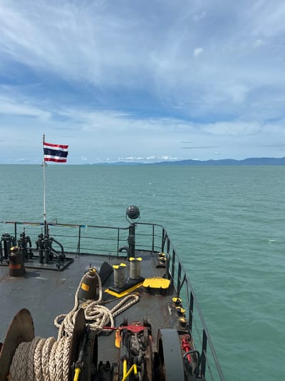 An overhead view from the deck of a ferry shows the turquoise ocean and a cloudy sky. In the foreground, a coil of thick rope is wrapped around a winch. Further back on the deck, bollards, valves, and a spotlight are visible. The Thai flag flies from a flagpole on the port side of the ferry. In the distance, a chain of islands can be seen rising from the ocean. The scene is peaceful and serene.