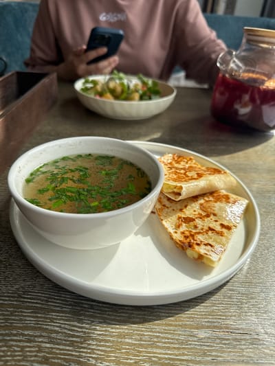 This media file captures a bright, sunlit meal scene set on a textured wooden table in Павловка, Russia. The central focus is a white round plate holding two main food items. On one side of the plate is a small, pristine white bowl filled with a clear broth soup, richly garnished with fresh green chives floating on the surface. Next to the soup, also on the white plate, are two pieces of a golden-brown, grilled flatbread, possibly a cheese quesadilla, folded into triangles, with melted cheese peeking out.
To the left of the main plate, a person is partially visible. She is wearing a light pink long-sleeved top and blue jeans. Her hand is holding a dark blue smartphone, suggesting she is interacting with it during the meal. Another white bowl, containing a fresh salad with various green leaves and possibly other ingredients, is partially visible closer to the person. Above the person's hand, a clear glass pitcher with a wooden lid, filled with a deep red beverage (possibly juice or kompot), is either being held or has recently been placed on the table.
The strong natural light indicates it is daytime, with distinct shadows cast on the table and the items, suggesting a sunny day, likely morning or early afternoon. The wooden table surface displays a prominent grain, contributing to a casual, possibly outdoor or semi-outdoor, dining ambiance. No discernible text is visible on any objects or clothing in the image.