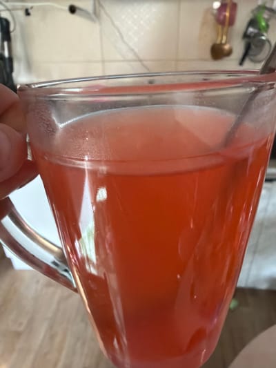 The image is a close-up, eye-level shot of a clear glass mug filled with a red-colored liquid, likely a beverage such as juice or tea. A spoon is partially submerged in the liquid, suggesting it has been stirred or is being used to drink. The mug is held by a hand, only partially visible on the left side of the frame, with fingers gripping the handle. The background is out of focus but appears to be an indoor setting with what looks like a kitchen counter and some hanging objects. The lighting is bright, creating reflections on the glass mug. The overall scene is simple and domestic, with no identifiable landmarks or specific urban features that would allow for pinpointing a location within Lopatino, Russia.