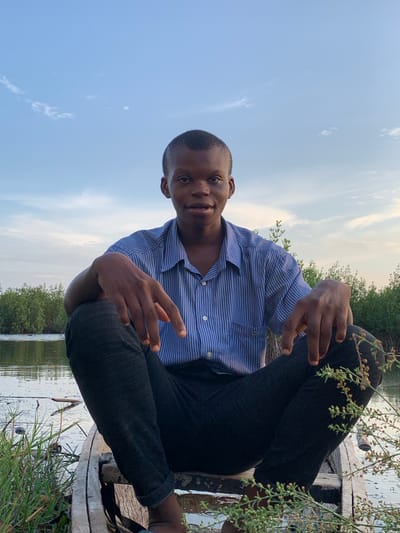A young Black man is the central subject of this outdoor portrait, seated and looking directly at the camera with a pleasant, subtle smile. He is wearing a blue and white striped short-sleeved collared shirt and dark trousers. His hands are resting casually on his bent knees. He appears to be sitting on a wooden structure, possibly a boat or a platform, positioned at the edge of a body of water. Green leafy branches are visible in the immediate foreground, particularly on the right side of the frame.

The background features calm water that reflects the light blue sky, indicating a clear day with soft, diffused sunlight, suggesting either early morning or late afternoon. Across the water, a dense line of green trees or bushes forms the horizon. The setting evokes a serene, natural environment, consistent with a river, creek, or coastal area, such as a mangrove ecosystem in Ibasa, Nigeria. No visible text is present in the image.