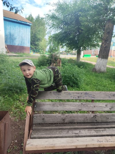 In Askiz, Russia, a young boy with a cap and camouflage clothing is climbing onto a wooden bench. He is positioned on all fours, with his left hand grasping the edge of the bench. The bench has graffiti scratched into its surface. The background features a blue wooden building, trees, and a grassy area with some foliage. The sky is cloudy with patches of blue.