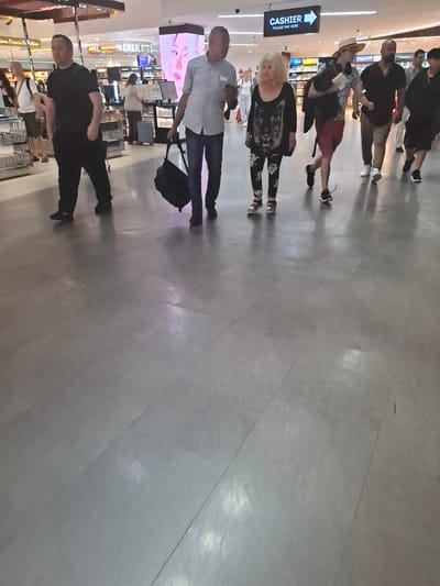 The image depicts a group of people walking through an indoor space, possibly an airport or a large shopping mall in Pendik, Turkey. In the foreground, on the left, a man wearing a black t-shirt and black pants is walking. To his right, a man in a light blue checkered shirt and dark pants is carrying a black duffel bag. Further to the right, a woman with blonde hair is wearing a black top with a tiger print and patterned leggings, walking alongside a man in a straw hat and patterned shirt. Behind them, a man in a dark t-shirt and khaki pants is walking, followed by another man in a black t-shirt. Several other individuals are visible in the background. The floor is a polished grey material. Overhead, a sign reads "CASHIER PLEASE PAY HERE" with an arrow pointing to the right. The environment appears to be a commercial area with displays and lighting consistent with retail spaces.