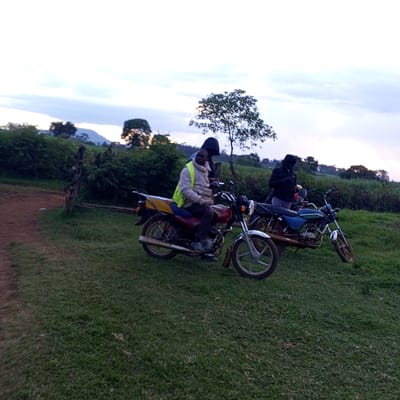 The image captures a scene in the Keyian ward of Kenya, featuring two motorcycles parked on a grassy area. A man wearing a yellow vest is seated on the red motorcycle. Another person, cloaked in dark clothing with a hood up, stands near the second, blue motorcycle. In the background, there's lush green vegetation, a tree, and a glimpse of a hill or mountain. The sky is overcast, suggesting either dusk or dawn.