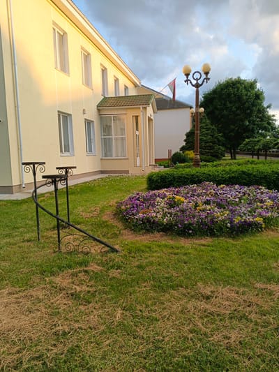 The image shows the exterior of a light yellow building in Baranavichy, Belarus. The building has several rectangular windows and a covered entrance with a green tiled roof. In front of the building, there is a well-maintained garden area with a large flower bed filled with purple, yellow, and white flowers. A lamppost with two globe-shaped lights stands next to the flower bed. To the left of the flower bed, there is a black metal rack with three circular platforms, and a decorative black metal railing with scrollwork. The grass in the foreground appears dry and patchy in some areas. A flag is visible on a pole attached to the building. The sky is cloudy.