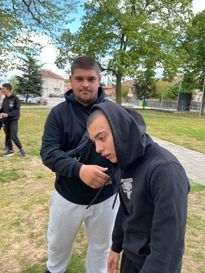 Two young men are photographed in an outdoor setting. The first man, positioned on the left, has short dark hair and a short beard. He is wearing a black hoodie. The second man is directly beside the first one. He has short dark hair, braces, and is also wearing a black hoodie with a graphic on the sleeve, along with light-gray sweatpants. The two men are standing together. In the background, there are at least two young men walking. The scene takes place in a grassy area, with trees and buildings visible in the background. The photo was taken in Karavelovo, Bulgaria.