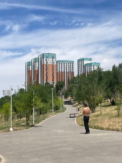 A man, shirtless and wearing dark pants and a baseball cap, stands facing away from the camera on a paved path in a park-like setting. The path curves to the left and ascends a gentle slope. To the right of the path, dry grass and scattered trees dominate the landscape. In the background, several modern, multi-story residential buildings are visible, characterized by their colorful facades featuring orange and green accents. The sky above is partly cloudy with a mix of blue sky and wispy white clouds. Park amenities, such as benches and lampposts, are also present along the path. The scene appears to be in the city of Almaty, Kazakhstan, specifically near the foot of the Alatau mountain range, a common area for residential development and recreational spaces.