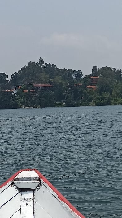 The image presents a scene from a body of water in Bwishyura, Rwanda, with a populated hillside in the background. In the foreground, the weathered white bow of a wooden boat is visible, featuring a distinct red trim along its top edge, suggesting the vantage point is from aboard the vessel. The midground is dominated by the expansive surface of the lake, displaying a deep blue-grey hue with gentle ripples. In the background, a large, lush green hill rises from the opposite shore, densely covered with trees and varied vegetation. Multiple multi-story buildings with prominent orange-red tiled roofs are interspersed among the trees on the hillside. The sky above is a uniform, light grey, indicating an overcast or hazy atmospheric condition.