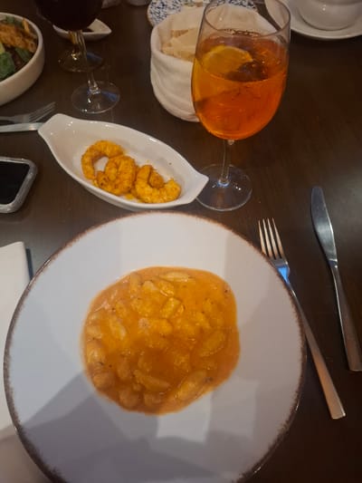 The image shows a restaurant table setting with a plate of gnocchi in an orange sauce, a small dish of spiced shrimp, a glass of Aperol spritz with a lemon slice, bread, a salad, a fork, and a knife. A mobile phone is also on the table. The location is inferred to be in Jesús del Monte, Mexico.